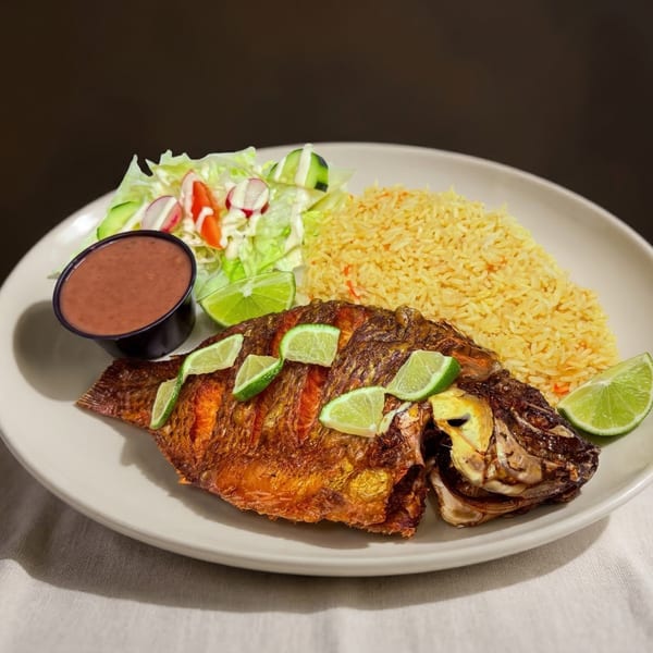 Whole fried tilapia with lime slices, yellow rice, a side salad, and red sauce on a white plate.