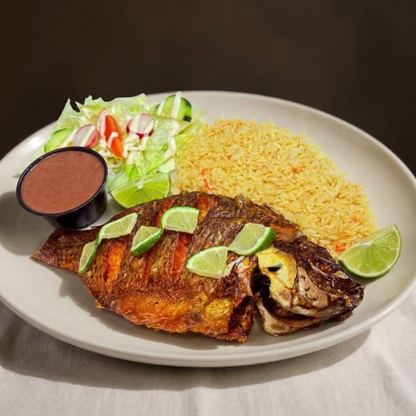 Whole fried fish with lime slices, yellow rice, a side salad, and red sauce on a white plate.