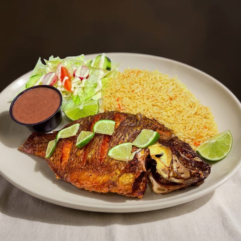 Whole fried fish with lime slices, yellow rice, a side salad, and red sauce on a white plate.