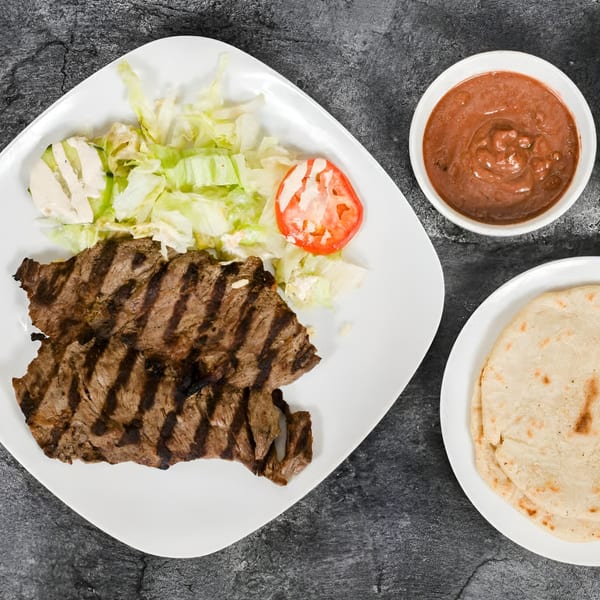 Grilled steak with a side salad, red sauce, and tortillas on a dark gray surface.