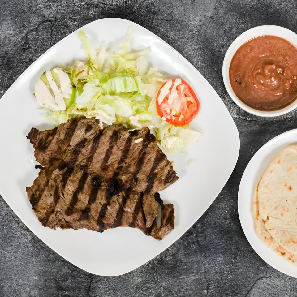 Grilled steak with lettuce, tomato, and cheese on a white plate, with salsa and tortillas.